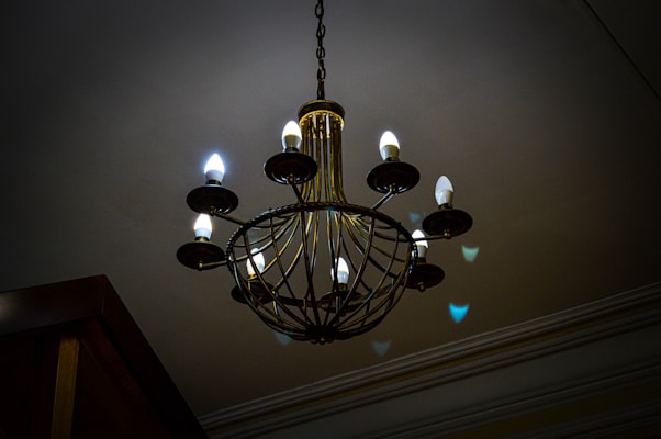 A large, ornate chandelier with a series of candle-like light bulbs arranged in a circular pattern is suspended from the ceiling. The lighting gives off a warm and dim ambiance, casting soft shadows and reflections on the ceiling and walls. The chandelier features intricate metalwork in a vintage style.
