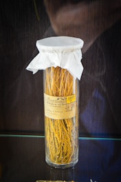 A tall glass jar with yellow plant material inside, sealed with a white paper cover and labeled with handwritten text.