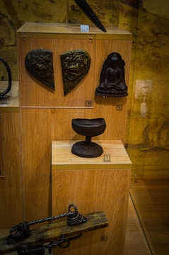 Various historical artifacts displayed in a wooden case, including a heart-shaped metal piece with intricate designs, a small sculpture of a seated figure, a rounded pedestal-like object, and an antique wooden tool with twisted metal components.