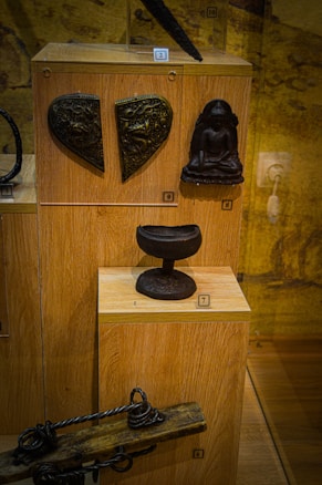 Various historical artifacts displayed in a wooden case, including a heart-shaped metal piece with intricate designs, a small sculpture of a seated figure, a rounded pedestal-like object, and an antique wooden tool with twisted metal components.