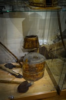 The image depicts a display of vintage wooden tools and equipment inside a glass case. There is a cylindrical wooden object with a hole in the center, possibly an old grinding tool or part of a mill. A square wooden container and several shovel-like tools with wooden handles are positioned nearby. The background shows a faint wall with some indistinct elements.