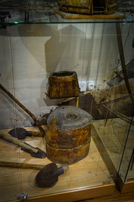 The image depicts a display of vintage wooden tools and equipment inside a glass case. There is a cylindrical wooden object with a hole in the center, possibly an old grinding tool or part of a mill. A square wooden container and several shovel-like tools with wooden handles are positioned nearby. The background shows a faint wall with some indistinct elements.