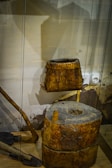 Vintage wooden tools are displayed in a glass case, featuring a large cylindrical piece resembling a mortar with a hollowed-out section on top. Adjacent to it is a wooden object that might have been used for carrying or storage. The background showcases a slightly blurred and abstract design, which gives a rustic feel to the setup.