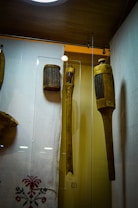 A display case contains three wooden objects, each designed with vertical slats or grooves, suggesting percussion instruments or similar artifacts. The objects are mounted on a light-colored background with fabric below, embroidered with a traditional floral pattern. Lighting highlights the warm tones of the wood, with a modern ceiling light fixture in view.