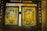 Two ornate golden religious icons are set within a wooden frame. The icons are richly decorated with detailed engravings, featuring religious figures surrounded by intricate patterns and floral motifs. The woodwork surrounding the icons is dark and rustic, contrasting with the vivid gold.