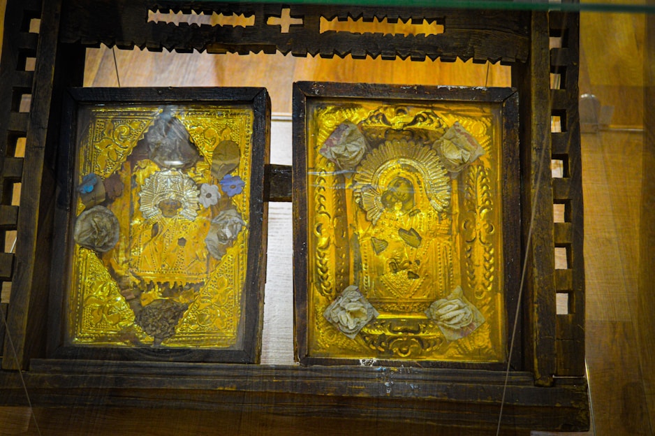 Two ornate golden religious icons are set within a wooden frame. The icons are richly decorated with detailed engravings, featuring religious figures surrounded by intricate patterns and floral motifs. The woodwork surrounding the icons is dark and rustic, contrasting with the vivid gold.
