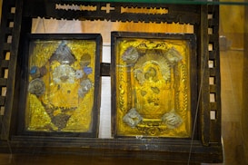 Two ornate golden religious icons are set within a wooden frame. The icons are richly decorated with detailed engravings, featuring religious figures surrounded by intricate patterns and floral motifs. The woodwork surrounding the icons is dark and rustic, contrasting with the vivid gold.