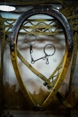 An old wooden and metal horse collar is displayed, with intricate rope work surrounding it. The metal piece at the top appears rusted, and the collar's shape is slightly oval. Behind it, a textured background adds depth to the scene.