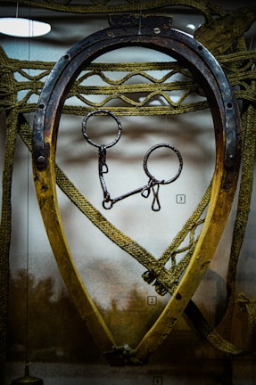 An old wooden and metal horse collar is displayed, with intricate rope work surrounding it. The metal piece at the top appears rusted, and the collar's shape is slightly oval. Behind it, a textured background adds depth to the scene.