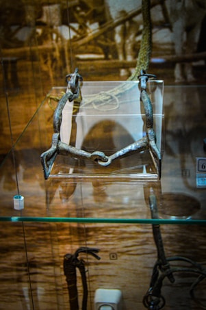 A metal object with a horse bit design is displayed in a glass case. The background features a historical sepia-toned photo that adds context to the antiquity of the object. The display environment suggests a museum or exhibit setting.