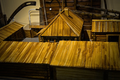Wooden model buildings featuring log cabin construction and prominent plank roofs dominate the scene. The craftsmanship displays detailed joints and fittings that reflect traditional architecture. The structures are arranged in a way that suggests a small village or settlement.