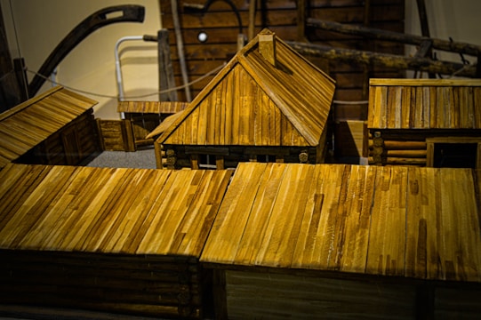 Wooden model buildings featuring log cabin construction and prominent plank roofs dominate the scene. The craftsmanship displays detailed joints and fittings that reflect traditional architecture. The structures are arranged in a way that suggests a small village or settlement.