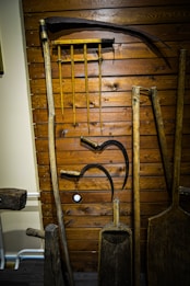 A collection of vintage wooden farming tools hangs on a wooden wall. The tools include sickles, a rake with wooden handles, and other implements. The wood shows signs of age and use, giving a rustic, historical texture.