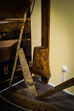 An old wooden agricultural or construction tool is leaning against a wall, paired with a rustic wooden object hanging on the wall. The setup suggests a historical or traditional setting, possibly part of a museum display. The floor is wooden, and there's an electrical outlet on the beige wall.