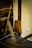 An old wooden agricultural or construction tool is leaning against a wall, paired with a rustic wooden object hanging on the wall. The setup suggests a historical or traditional setting, possibly part of a museum display. The floor is wooden, and there's an electrical outlet on the beige wall.