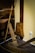An old wooden agricultural or construction tool is leaning against a wall, paired with a rustic wooden object hanging on the wall. The setup suggests a historical or traditional setting, possibly part of a museum display. The floor is wooden, and there's an electrical outlet on the beige wall.