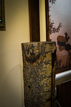 A textured wooden log with visible bark standing against a cream-colored wall. There is a framed artwork partially visible on the wall to the left and a painting depicting a person and foliage to the right. The log appears to be a decorative or artistic element in the interior setting.
