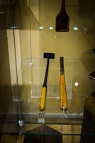 Two wooden-handled tools encased in a glass display, with distinct dark metallic working ends. One appears to be a small hammer and the other a chisel. A separate wooden tool with a wider head hangs above them. The background is beige, and there are reflection spots from the lights on the glass.