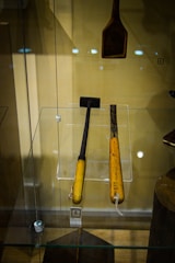 Two wooden-handled tools encased in a glass display, with distinct dark metallic working ends. One appears to be a small hammer and the other a chisel. A separate wooden tool with a wider head hangs above them. The background is beige, and there are reflection spots from the lights on the glass.