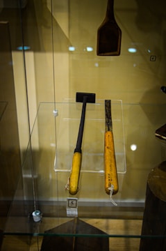 Two wooden-handled tools encased in a glass display, with distinct dark metallic working ends. One appears to be a small hammer and the other a chisel. A separate wooden tool with a wider head hangs above them. The background is beige, and there are reflection spots from the lights on the glass.