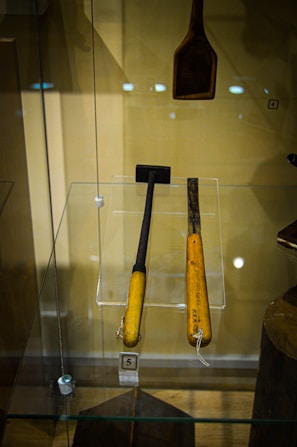 Two wooden-handled tools encased in a glass display, with distinct dark metallic working ends. One appears to be a small hammer and the other a chisel. A separate wooden tool with a wider head hangs above them. The background is beige, and there are reflection spots from the lights on the glass.