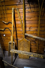 A variety of old wooden tools are displayed against a wooden wall. The tools include large paddles and wooden handles, possibly used for agricultural or manual tasks. The wood has a weathered look, indicating age and use.