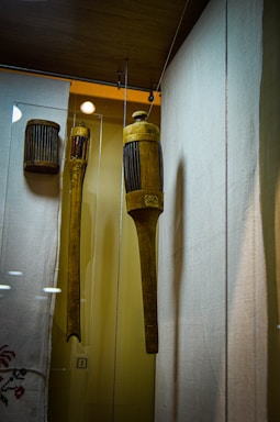 A display of wooden historical objects encased in glass. The main object is a cylindrical, intricately carved wooden tool with vertical grooves, hanging alongside another similar object. The background includes a fabric with delicate embroidery, and the setting appears to be a museum.