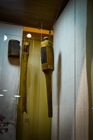 A display of wooden historical objects encased in glass. The main object is a cylindrical, intricately carved wooden tool with vertical grooves, hanging alongside another similar object. The background includes a fabric with delicate embroidery, and the setting appears to be a museum.