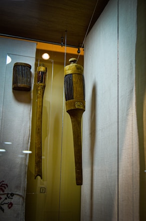A display of wooden historical objects encased in glass. The main object is a cylindrical, intricately carved wooden tool with vertical grooves, hanging alongside another similar object. The background includes a fabric with delicate embroidery, and the setting appears to be a museum.