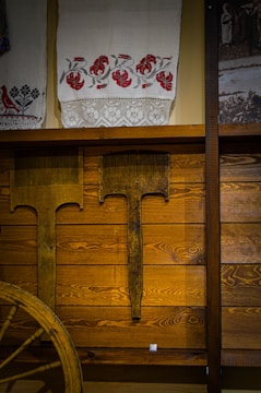 A rustic interior featuring wooden paneling and a display of traditional textiles. The textiles are adorned with embroidered red floral patterns and lace details. Antique farming tools are mounted on the wall beneath, and part of an old wooden wagon wheel is visible, adding to the vintage theme.