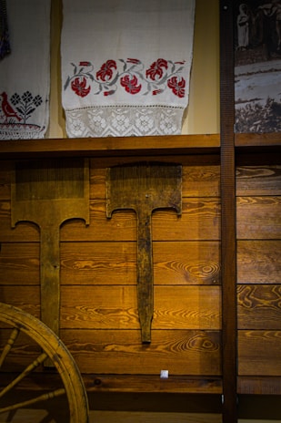 A rustic interior featuring wooden paneling and a display of traditional textiles. The textiles are adorned with embroidered red floral patterns and lace details. Antique farming tools are mounted on the wall beneath, and part of an old wooden wagon wheel is visible, adding to the vintage theme.