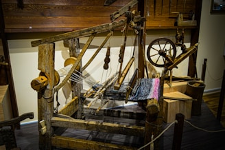 A rustic wooden loom with an intricate setup of pulleys, gears, and threads, partially weaving a multi-colored textile. The loom is surrounded by wooden tools and components reminiscent of traditional weaving techniques. The wooden floor and walls enhance the old-fashioned, handcrafted aesthetic.