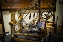 A rustic wooden loom with an intricate setup of pulleys, gears, and threads, partially weaving a multi-colored textile. The loom is surrounded by wooden tools and components reminiscent of traditional weaving techniques. The wooden floor and walls enhance the old-fashioned, handcrafted aesthetic.