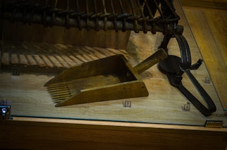 A vintage wooden and metal tool displayed on a wooden surface, alongside a few other metal implements. The wood appears aged, and the metal is slightly rusted, indicating historical use. Labels with numbers are attached, suggesting a museum or collection display.