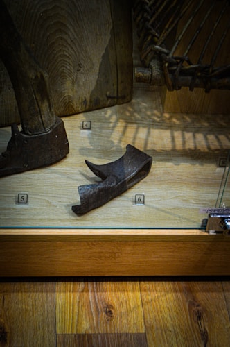 A museum exhibit displaying a collection of ancient tools or artifacts. The central focus is a wooden or metal implement placed on a wooden floor within a transparent case, with small numbered labels nearby.