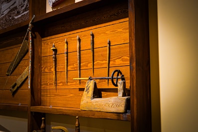 A wooden display case houses several antique tools, including spindles, a hand drill, and other woodworking implements. The background consists of dark stained wooden planks, adding to the rustic and historical feel of the setup.