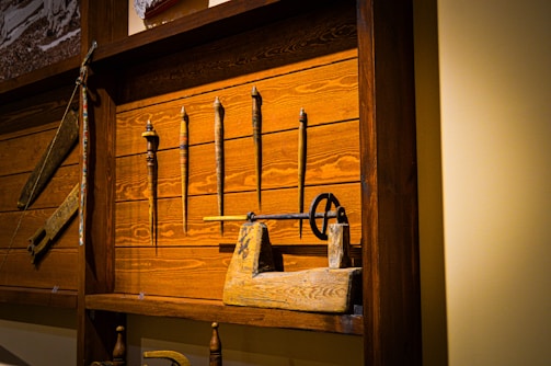 A wooden display case houses several antique tools, including spindles, a hand drill, and other woodworking implements. The background consists of dark stained wooden planks, adding to the rustic and historical feel of the setup.