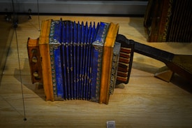 An antique accordion with a striking blue bellows and wooden ends adorned with intricate patterns sits on a wooden surface. The instrument displays signs of age and wear, with metal hardware securing the edges. Various buttons and keys are visible on its side, used for producing music.