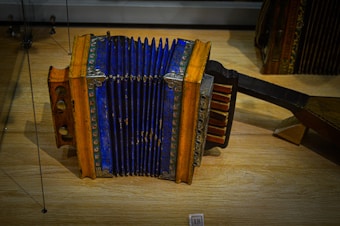An antique accordion with a striking blue bellows and wooden ends adorned with intricate patterns sits on a wooden surface. The instrument displays signs of age and wear, with metal hardware securing the edges. Various buttons and keys are visible on its side, used for producing music.