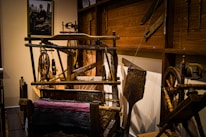 A rustic room displaying traditional weaving equipment, including spinning wheels and a wooden loom with threads. The walls are adorned with wooden panels, and a framed photograph is visible in the background. The scene conveys a sense of historical craftsmanship.