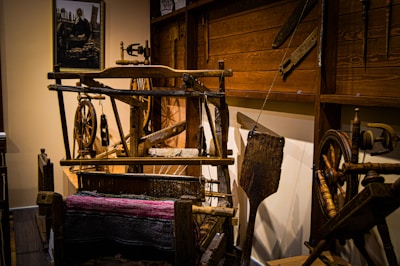 A rustic room displaying traditional weaving equipment, including spinning wheels and a wooden loom with threads. The walls are adorned with wooden panels, and a framed photograph is visible in the background. The scene conveys a sense of historical craftsmanship.