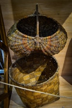 A rustic woven basket with a lid propped open, displaying intricate patterns and textures typical of handcrafts. The basket's materials appear natural and earthy, featuring a combination of tightly interwoven strips and a simple wooden structure. It's set against a wooden background, enhancing the traditional craftsmanship feel.