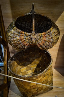 A rustic woven basket with a lid propped open, displaying intricate patterns and textures typical of handcrafts. The basket's materials appear natural and earthy, featuring a combination of tightly interwoven strips and a simple wooden structure. It's set against a wooden background, enhancing the traditional craftsmanship feel.