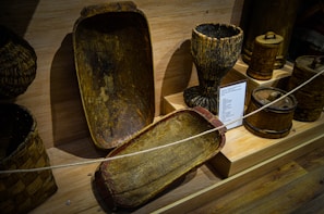 The image displays a collection of rustic wooden artifacts including large troughs, a goblet-like structure, and barrel-like containers. The items are arranged on a wooden surface and have an aged, rough texture with a natural finish.
