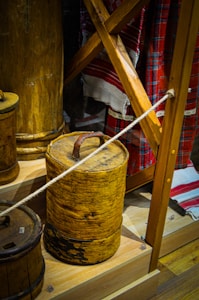 An image displays traditional wooden containers arranged on wooden shelves. The containers appear aged with a rustic patina, possessing handles on the top. The background includes fabric with red and white plaid and striped patterns. A rope barrier is present, suggesting a museum or exhibit setting.