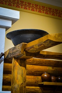 A rustic interior featuring wooden beams and logs with a decorative geometric pattern on the upper wall. A large black metal bowl is placed atop one of the beams, and there are several clay pots on a wooden shelf in the background.