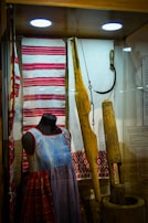 A display case features traditional textiles and clothing alongside carpentry tools. A mannequin sports a denim-like dress with red and plaid patterns. The textiles include red and white striped and embroidered designs. Wooden tools and a sickle are also present, hinting at historical agricultural practices.