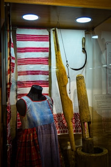 A display case features traditional textiles and clothing alongside carpentry tools. A mannequin sports a denim-like dress with red and plaid patterns. The textiles include red and white striped and embroidered designs. Wooden tools and a sickle are also present, hinting at historical agricultural practices.