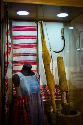A display case features traditional textiles and clothing alongside carpentry tools. A mannequin sports a denim-like dress with red and plaid patterns. The textiles include red and white striped and embroidered designs. Wooden tools and a sickle are also present, hinting at historical agricultural practices.