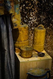 A display of intricately designed wooden containers and a fabric drape. The containers have detailed carvings and patterns, showcasing expert craftsmanship. The drape features a decorative design with blue and brown tones. The background has a textured appearance.